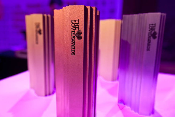 Four metallic trophies with The Love Awards logo stand upright on a pink-lit surface, reminiscent of Lovie Awards elegance. The trophies feature a modern, ridged design and vary in colour tones, set against a purple background.