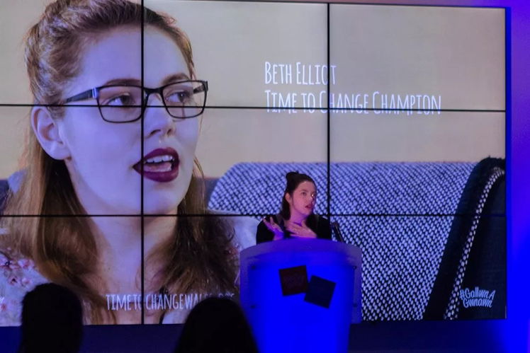 A woman stands at a lectern giving a speech at the #WeCanWeWill Launch Event, with a large screen behind her displaying a close-up of her face and the text: Beth Elliot, Time to Change Champion. The setting features blue lighting.