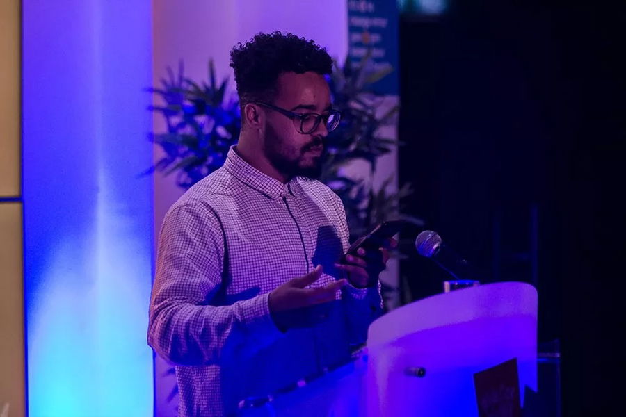A man with glasses and a checked shirt speaks at a lectern, holding a mobile at the #WeCanWeWill Launch Event. Blue and purple lighting set the mood, with a plant visible in the background.