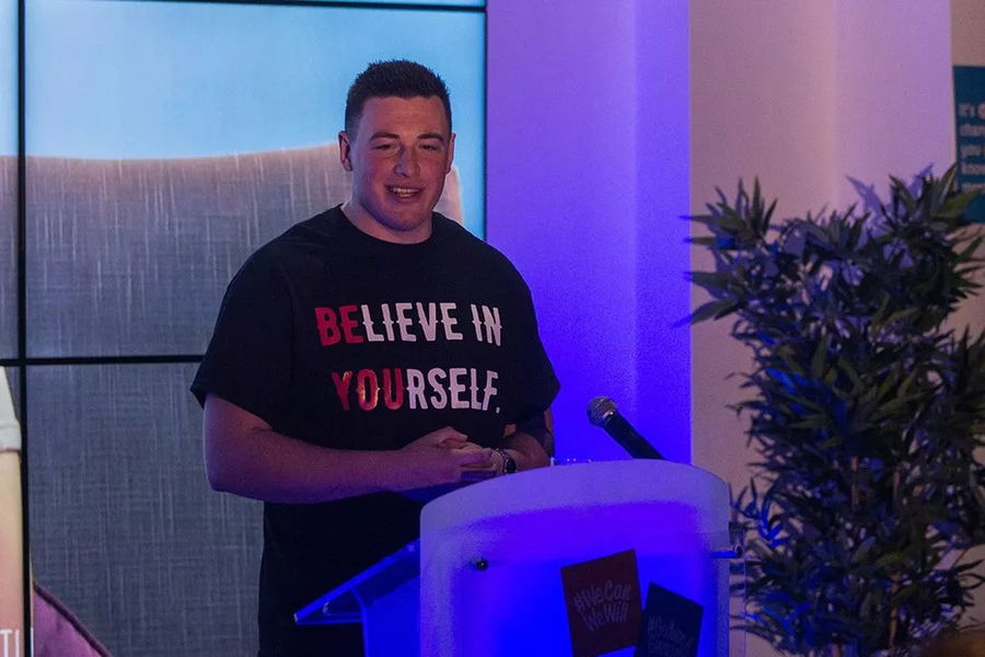 A person stands at a lectern, smiling, wearing a black T-shirt with the words “BELIEVE IN YOURSELF” in bold red and white letters at the #WeCanWeWill Launch Event. A plant and screens appear in the background.