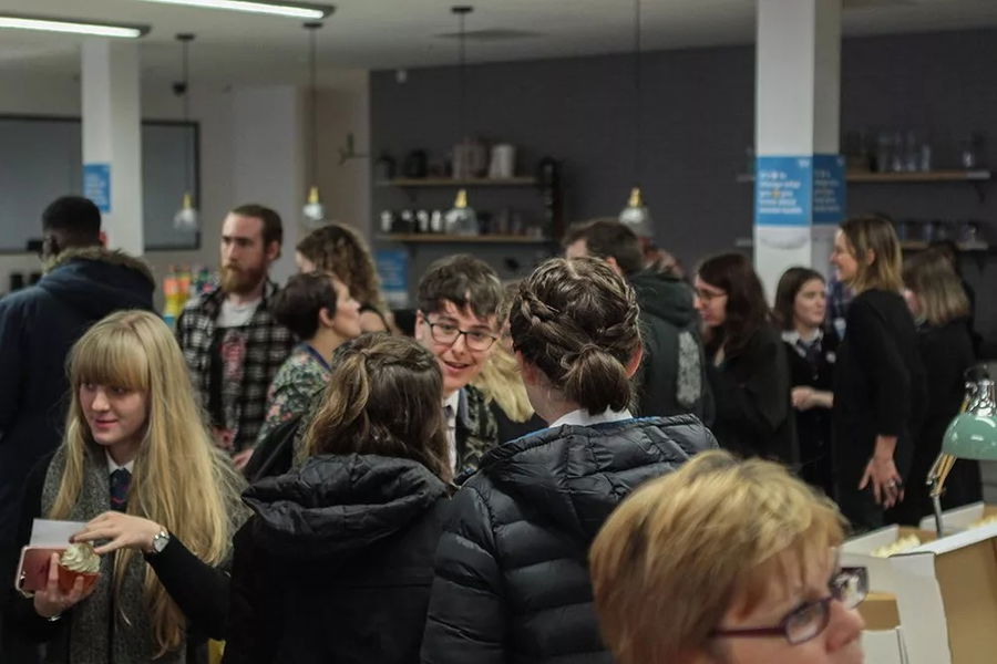 A group of people socialises indoors at what appears to be a casual #WeCanWeWill Launch Event, with some holding drinks or snacks. The room has warm lighting and shelves in the background.