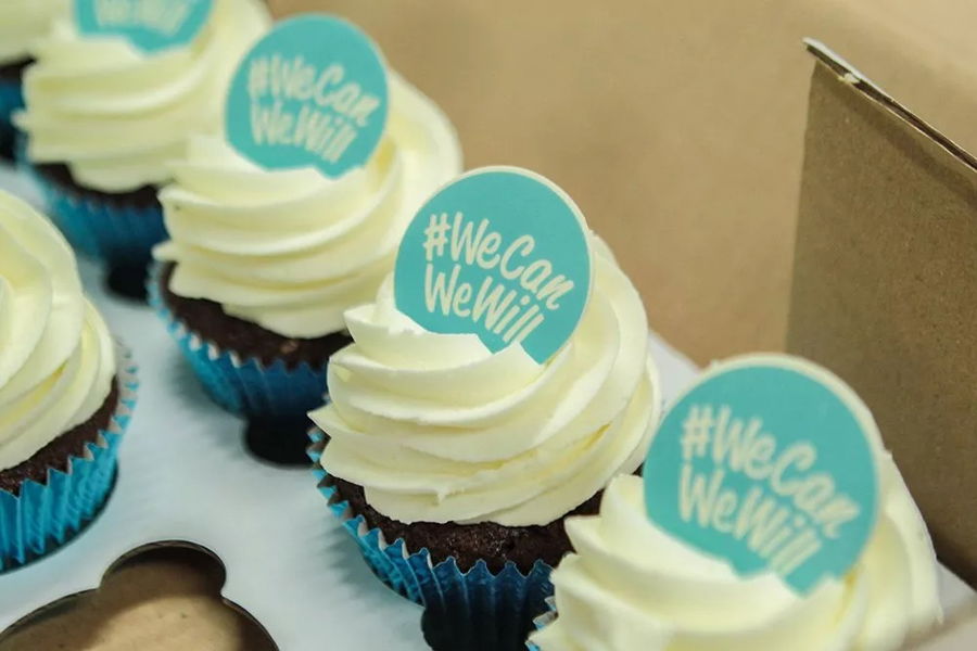 Chocolate cupcakes with white icing in blue cases, each topped with a #WeCanWeWill sign, are arranged in a box—perfect treats for the #WeCanWeWill Launch Event.