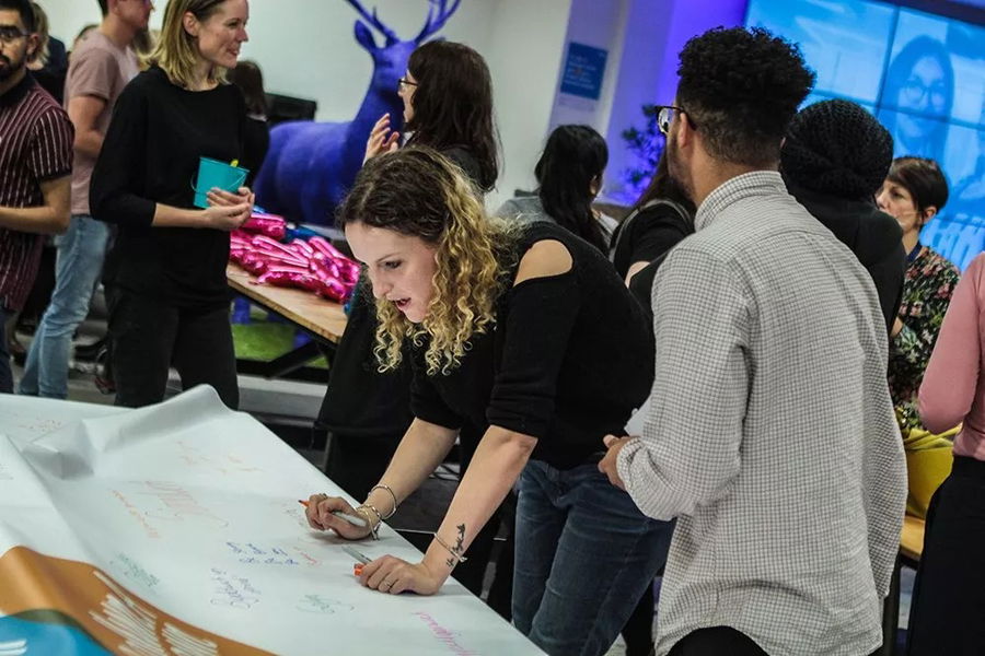 A woman with curly hair leans over a large sheet of paper, writing with a marker, whilst others stand and talk around her in a brightly lit, creative office space during the #WeCanWeWill Launch Event. A blue deer statue is visible in the background.
