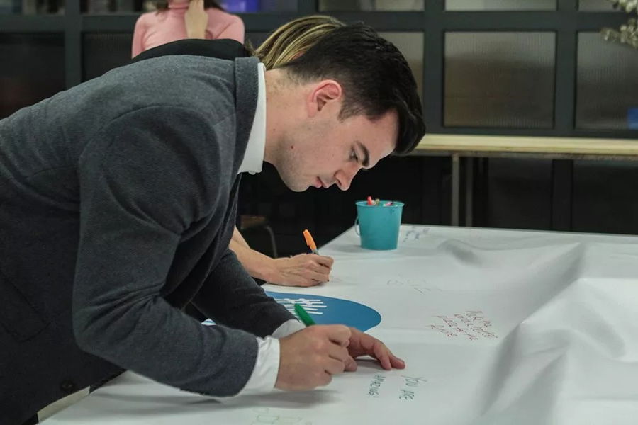 At the #WeCanWeWill Launch Event, a man in a grey suit jacket leans over a large sheet of paper, writing with a green marker. Another person beside him is also writing. There is a blue container with markers on the table.