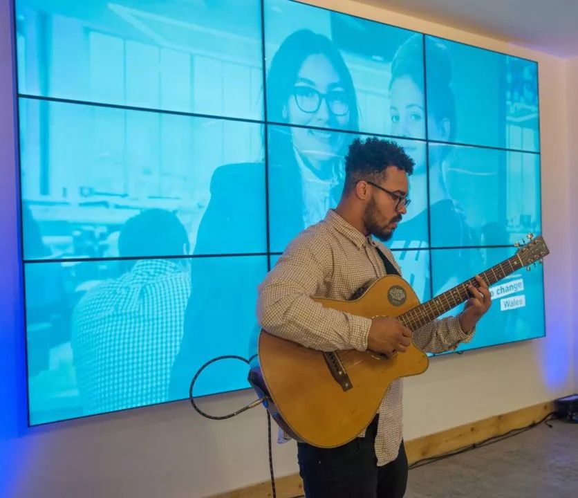 A man plays an acoustic guitar in front of a large, blue-tinted screen showing images of two women and an office setting. The indoor scene appears to be part of the #WeCanWeWill Launch Event or a similar presentation.