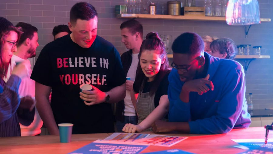 A group of young adults stand around a table, smiling and looking at papers at the #WeCanWeWill Launch Event. One person wears a shirt that says “BELIEVE IN YOURSELF” in this casual social gathering set in a modern kitchen or café.
