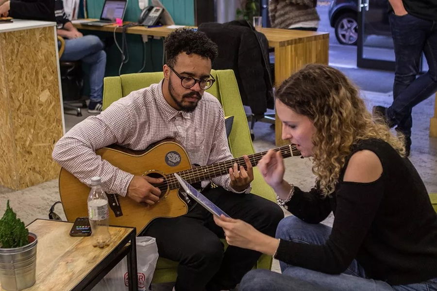 A man sits on a green chair playing an acoustic guitar while a woman beside him, possibly brainstorming for the #WeCanWeWill Launch Event, looks at a tablet and gestures in their modern, casual indoor office space.