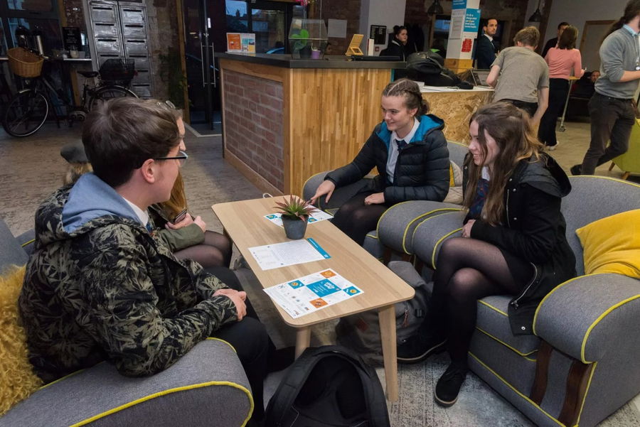 Four young people sit around a small table in a cosy lounge area at the #WeCanWeWill Launch Event, chatting and looking over papers and brochures. Other guests mingle near the counter, adding to the event’s casual and relaxed atmosphere.
