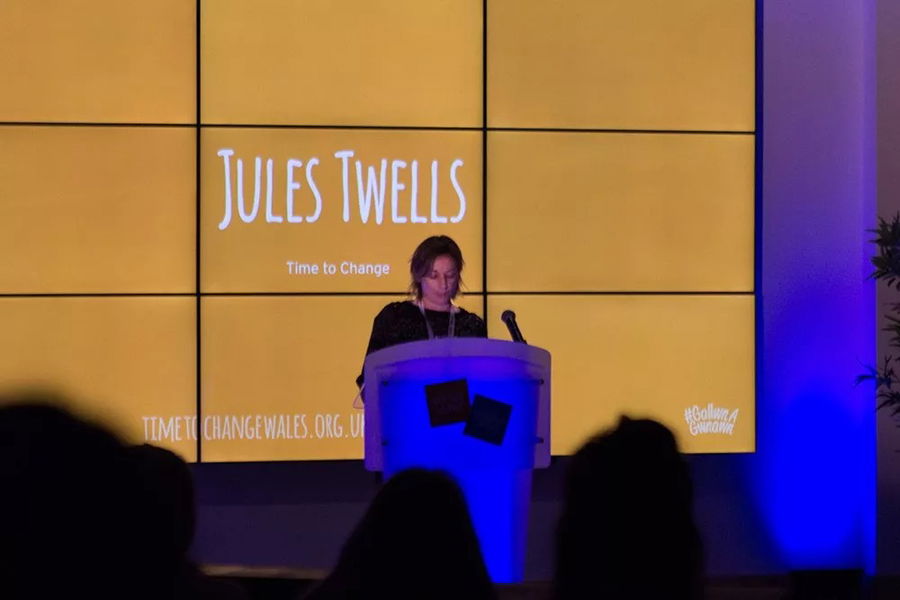 A woman stands at a lectern giving a speech in front of a large screen displaying the name Jules Twells and the phrase Time to Change at the #WeCanWeWill Launch Event. The background features a warm yellow colour.