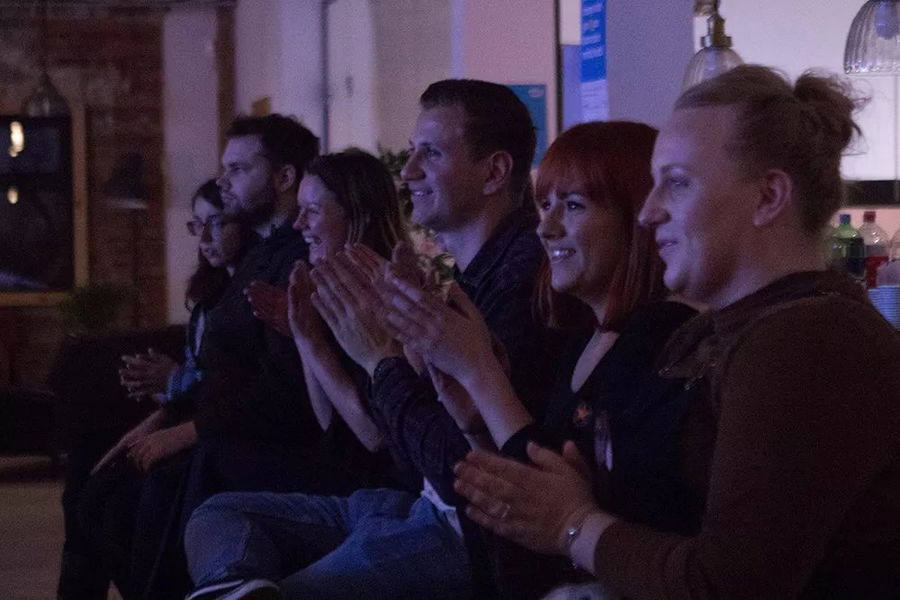 A group of people sitting in a row indoors, smiling and applauding whilst watching the #WeCanWeWill Launch Event, with dim lighting creating a cosy atmosphere.