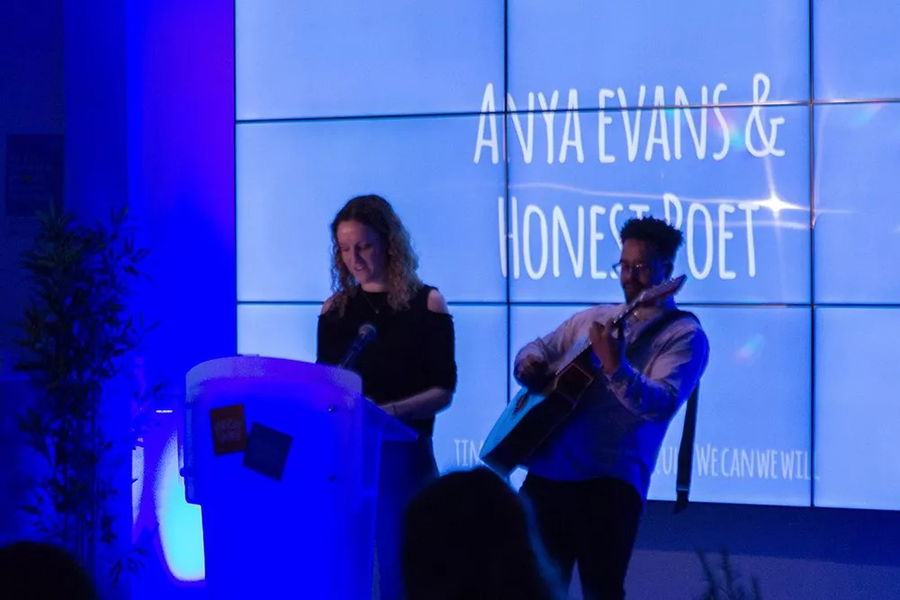 A woman stands at a lectern reading, while a man beside her plays guitar. They are performing in front of a large screen displaying Anya Evans & Honest Poet at the #WeCanWeWill Launch Event in a blue-lit room.