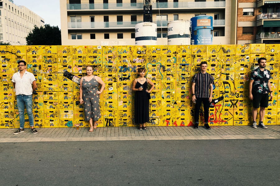Five people stand spaced apart in front of a yellow art wall covered in black doodles and graffiti, in an urban area with blocks of flats and building materials in the background.