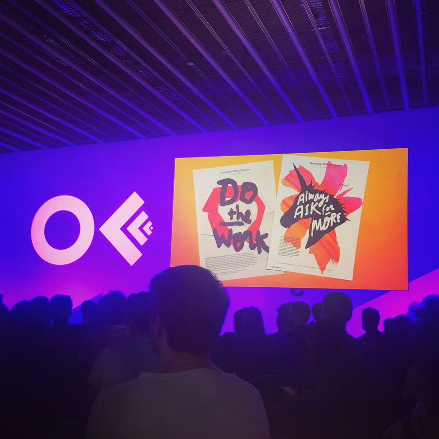 A group of people sit in a dark auditorium facing a screen displaying two colourful posters reading Do the Work and Always Ask for More. The background is lit with purple and the letters OE are projected on the wall.