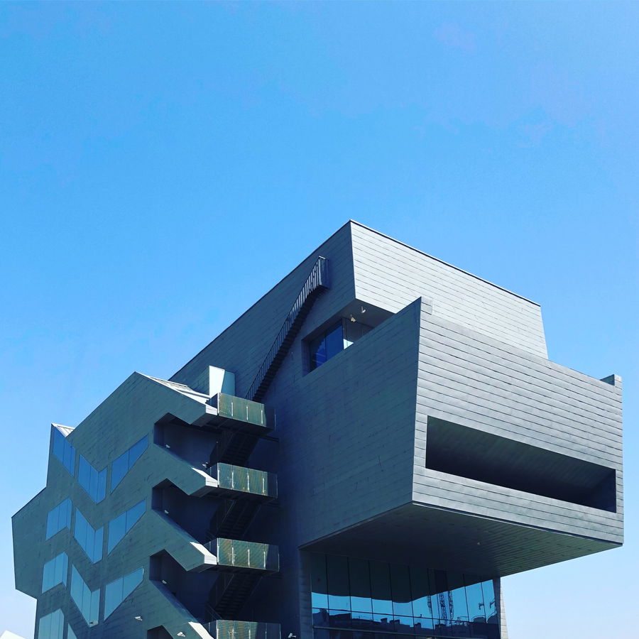 A modern building with angular, geometric architecture features glass windows and outdoor staircases, set against a clear blue sky.