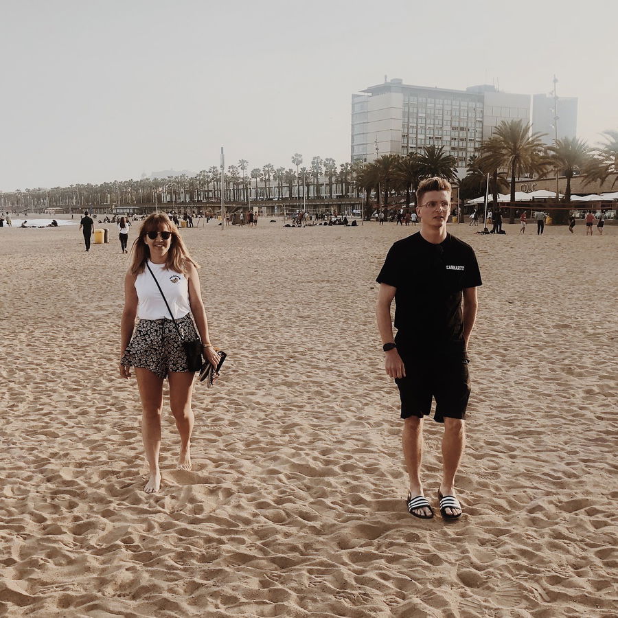 A woman and a man walk on a sandy beach with palm trees and a modern building in the background. The woman is barefoot, wearing a white top and patterned shorts; the man wears a black outfit with sliders. It's a sunny day.