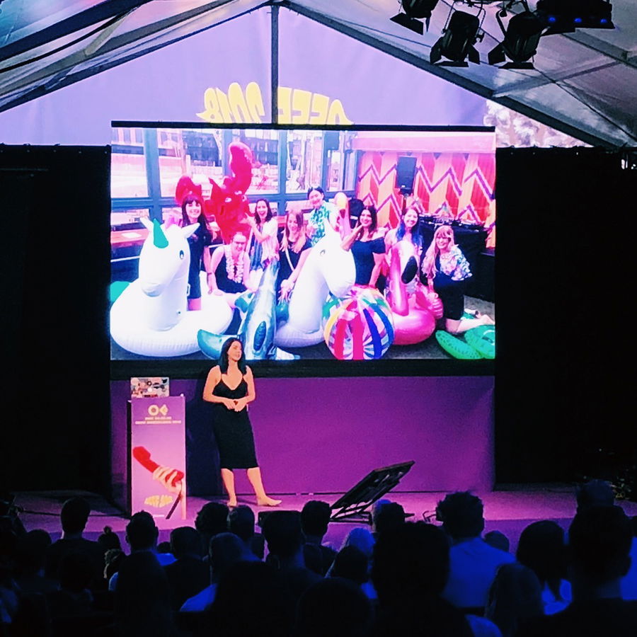 A woman stands on stage giving a presentation to an audience. Behind her is a large screen displaying a group photo of people with large inflatable pool toys, including unicorns and a lobster.