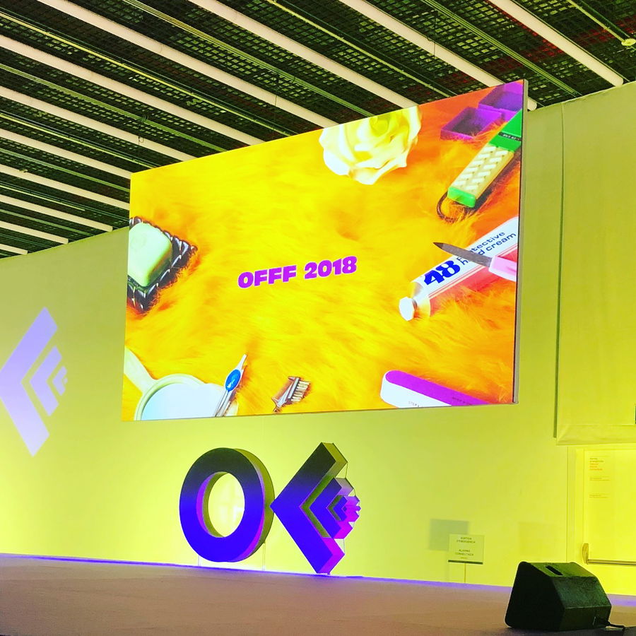 A large screen displays OFFF 2018 with colourful objects on a yellow background at an indoor event, with an OFFF logo sculpture on stage beneath the screen.