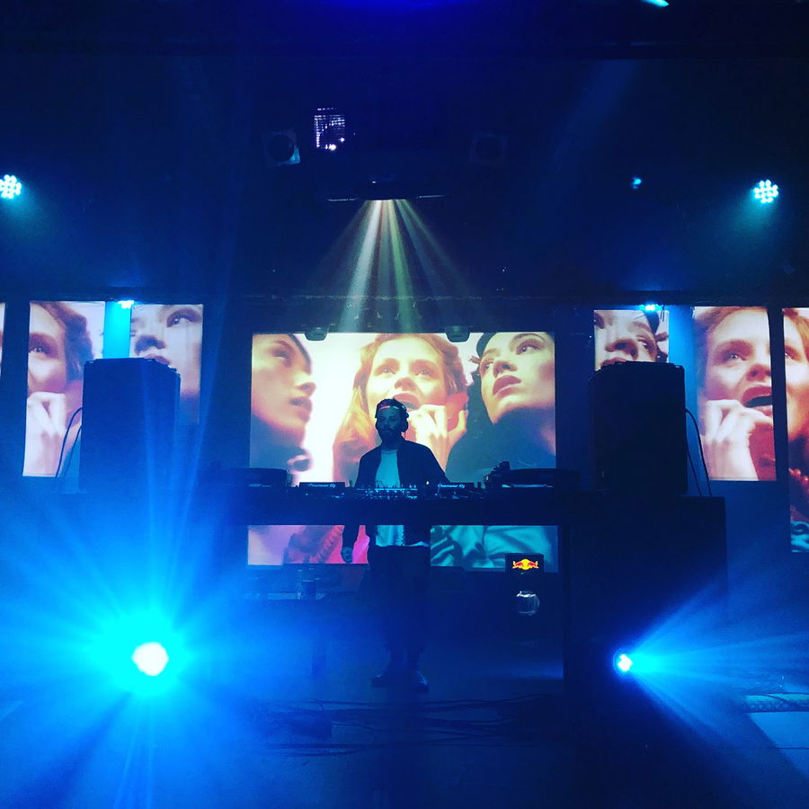 A DJ stands behind a booth on a dark stage, illuminated by bright blue lights. Large screens behind display images of a woman's face from different angles, creating a vibrant visual backdrop.