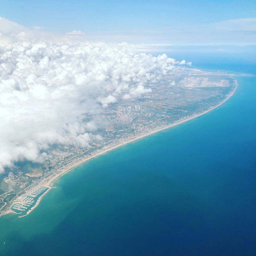 Aerial view of a coastline with blue sea, sandy beaches, a marina, and a city partially covered by white clouds under a clear sky.