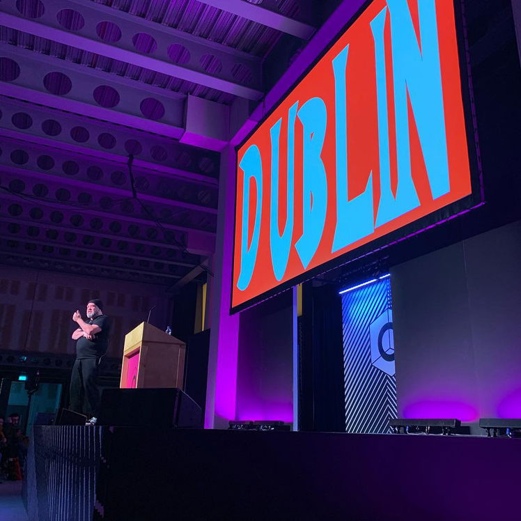 A speaker stands on stage next to a large screen displaying the word DUBLIN in bold, blue and red letters, under purple lighting at an indoor event or conference.