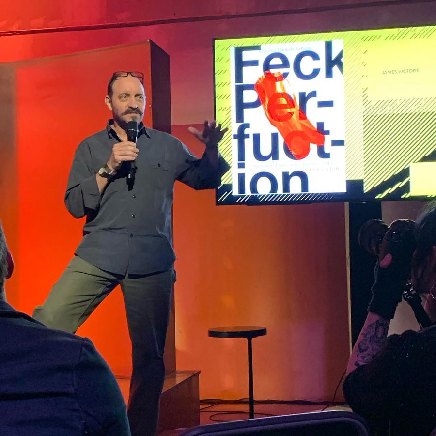 A man with a beard holds a microphone and speaks on stage next to a screen displaying the text Feck Perfection. The screen has abstract art and bold graphics. Several people are seated in the audience.