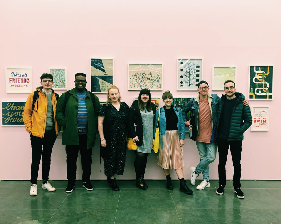 Seven people stand in a row, smiling at the camera in front of a light pink wall with colourful framed artwork. The group is casually dressed, and the setting appears to be an art gallery or exhibition.