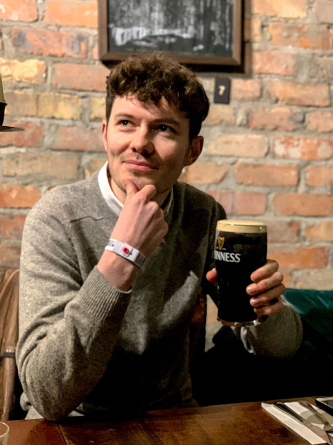 A young man in a grey jumper sits at a wooden table in front of a brick wall, holding a pint of Guinness and looking thoughtful with his hand on his chin.