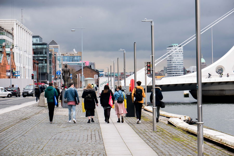 People walk along a cobblestone riverside path in a city, with modern buildings and a white bridge nearby. The sky is overcast, and the group walks away from the camera, some carrying rucksacks and bags.