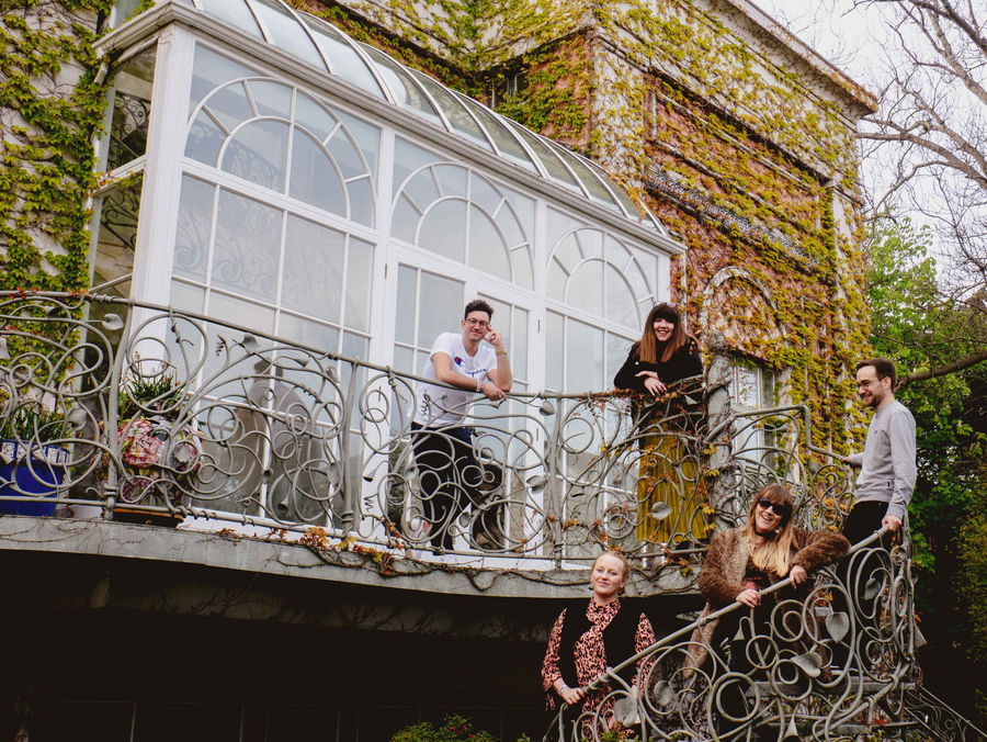 Five people stand and smile on an ornate iron balcony attached to a building covered in ivy. The building features large windows and an upper sunroom; trees and greenery surround the scene.