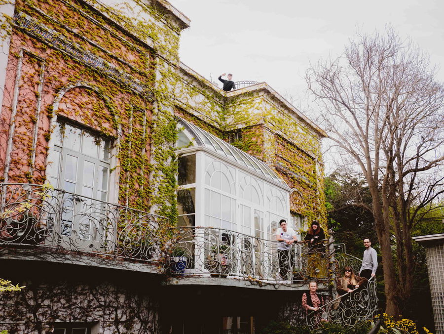 A group of people stand and sit on the ornate balcony of a large brick building covered in ivy, with one person waving from the rooftop and bare trees in the background.