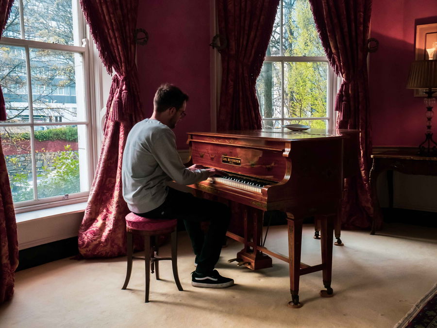 A person sits on a cushioned stool playing a wooden piano in a sunlit room with large windows, red curtains, and pink walls.