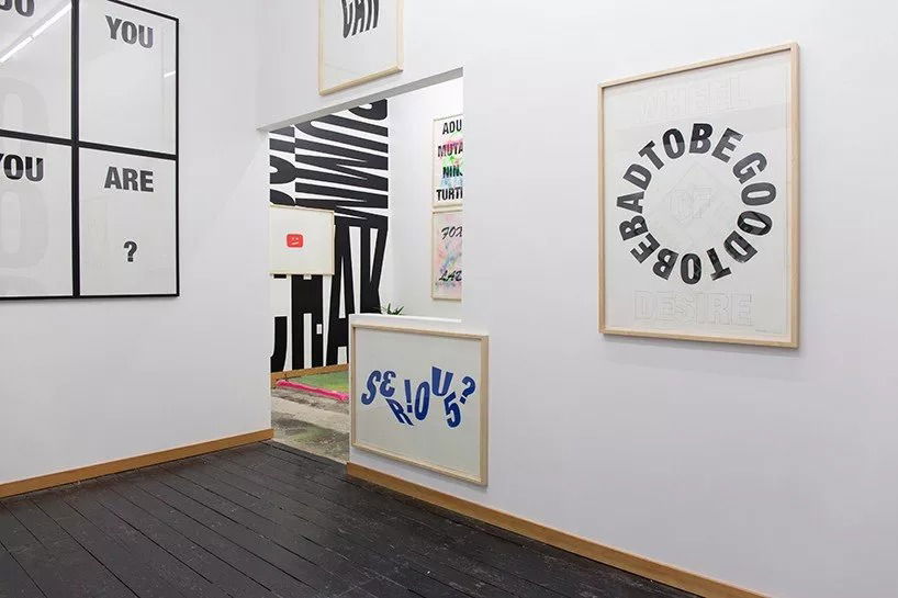 A modern art gallery with white walls displays various framed text-based artworks featuring bold, playful typography and questions. The floor is dark wood, and a colourful rug is visible in the adjoining room.