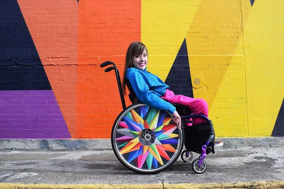 A woman in bright clothing smiles whilst sitting in a wheelchair with colourful, geometric designs on the wheels, in front of a vibrant mural with black, purple, orange, and yellow patterns.