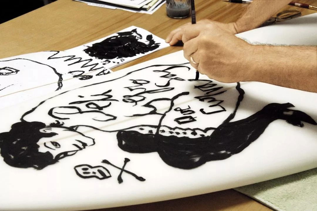 A person is painting a black, sketch-like illustration and stylised text onto a white surfboard. The drawing includes a portrait, a skull and crossbones, and various handwritten words. Papers and paint are on the table.