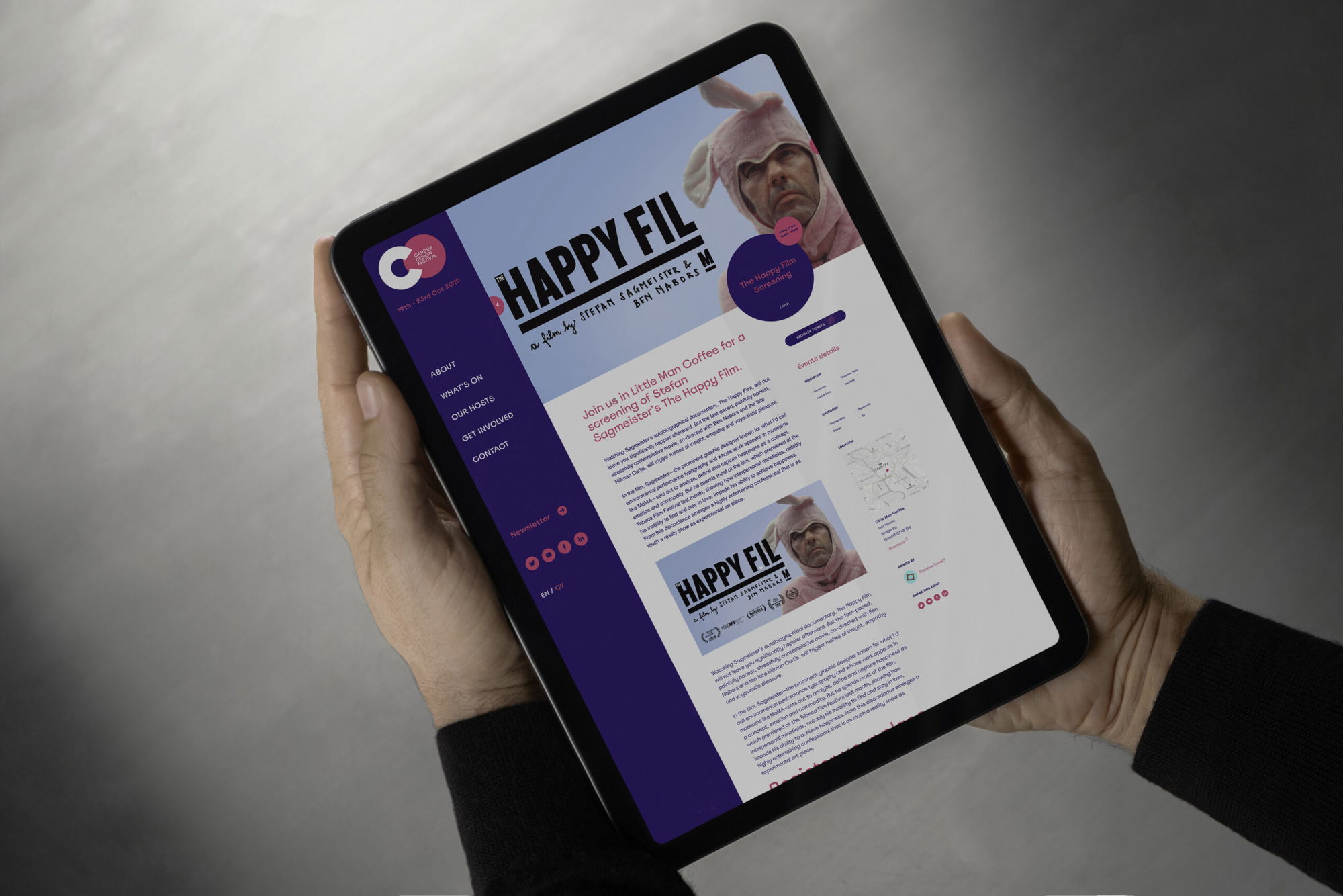 A person holds a tablet displaying a website about a programme called Happy Fil, featuring a man wearing a pink bunny hat. The website has a blue, purple, and pink colour scheme with various menu options on the left.