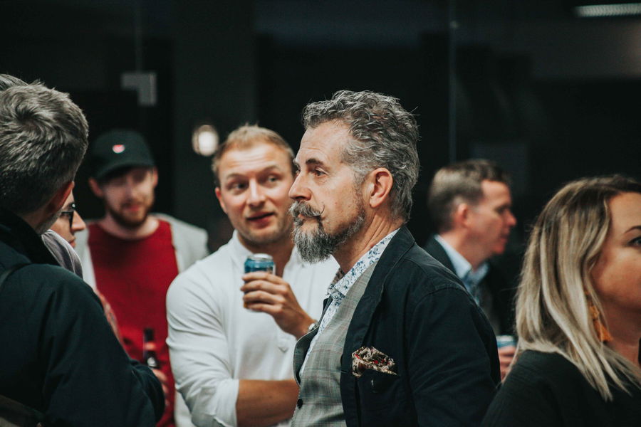 A group of people socialising indoors, some holding drinks. A man with grey hair and a beard stands in focus, while others engage in conversation around him. The atmosphere appears casual and lively.