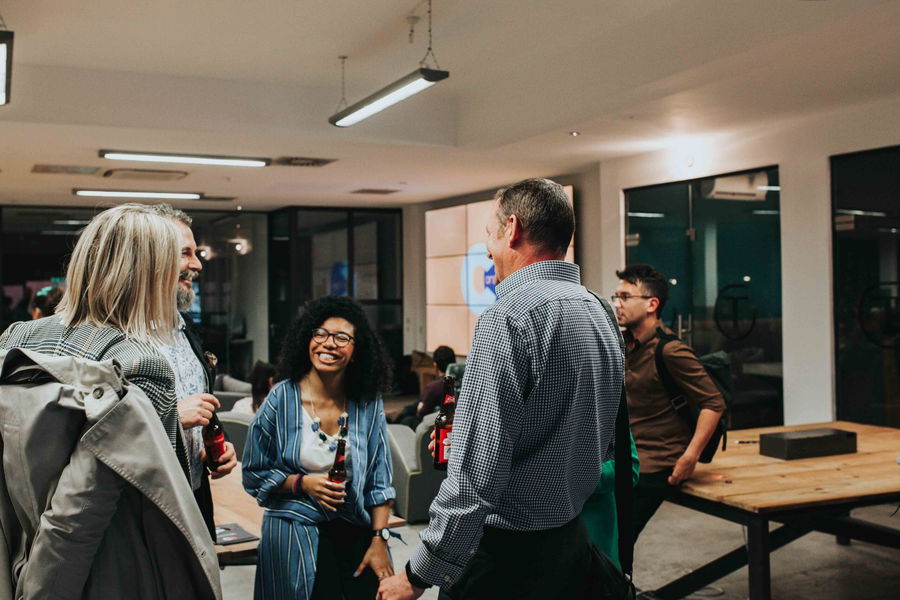 A group of people stand and chat in a modern office space, some holding bottled drinks. They appear to be enjoying a casual conversation, with one woman in the centre smiling brightly.
