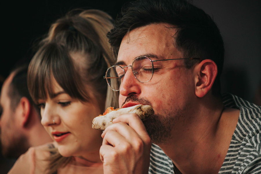 A man wearing glasses enjoys eating a slice of pizza with his eyes closed, while a woman with a fringe and a nose ring sits beside him, looking down. Both appear to be sitting together in an informal setting.