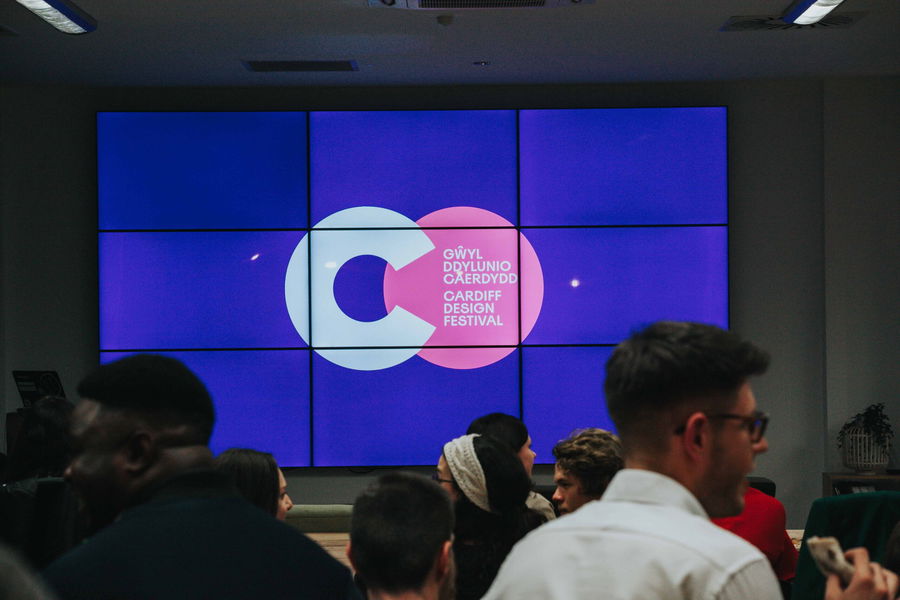 A group of people stand in front of a large screen displaying the Cardiff Design Festival logo, with overlapping blue and pink circles and text in Welsh and English.