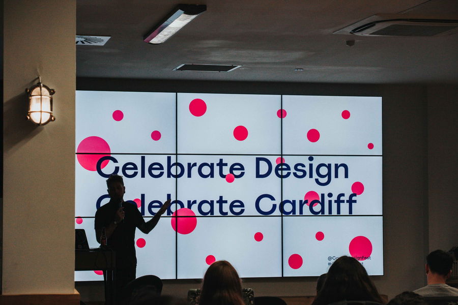 A person gives a presentation in front of a large screen displaying the text Celebrate Design Celebrate Cardiff with red dots on a white background. Audience members are seated in the foreground.