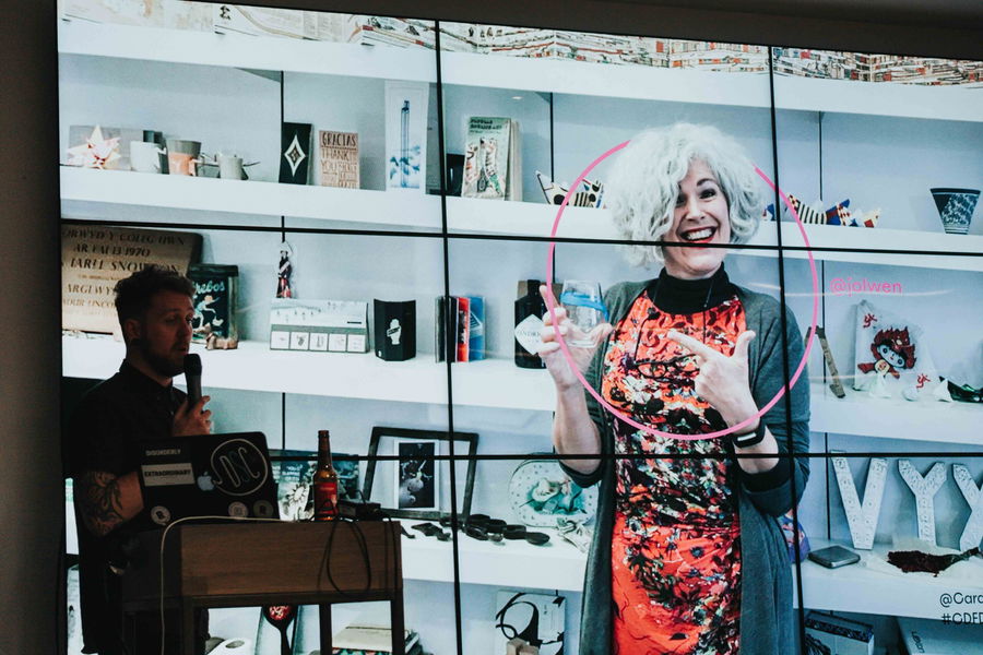 A person stands at a lectern with a microphone while a large screen behind them displays a smiling woman with curly grey hair, wearing a red dress and gesturing with her hands in front of shelves with various objects.