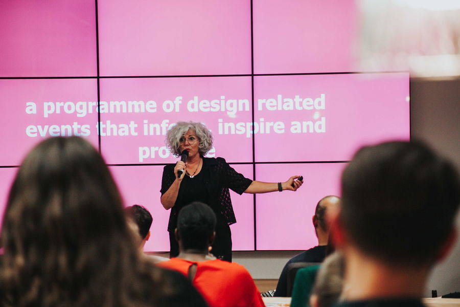A woman with curly grey hair speaks into a microphone on stage in front of a pink screen displaying white text to an audience. The text partially reads: a programme of design related events that inf... inspire and pro...