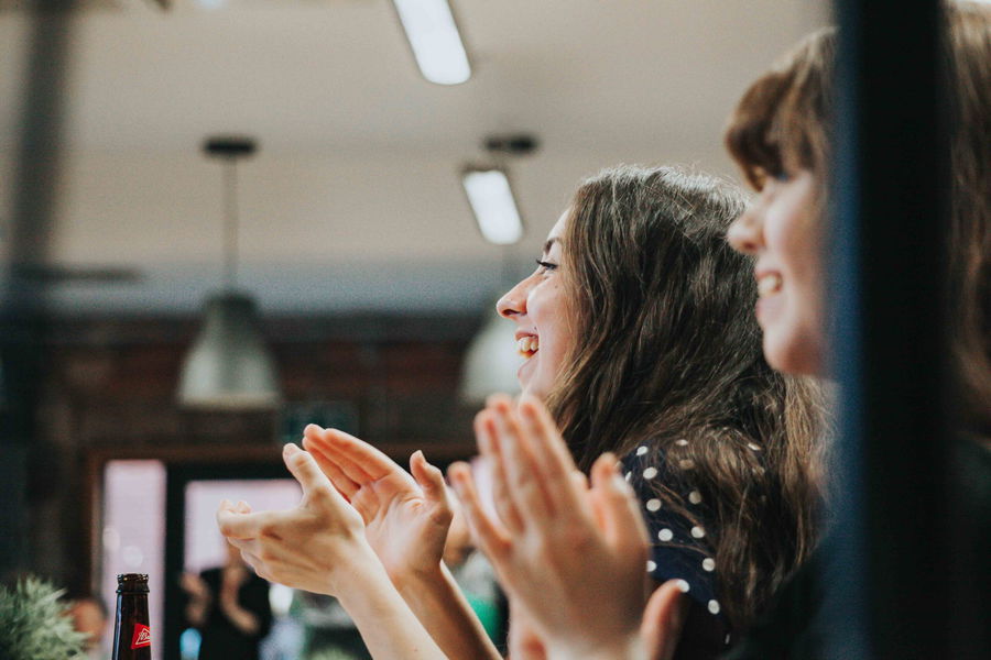 Two women are sitting side by side, smiling and clapping their hands in a brightly lit indoor setting. The atmosphere appears cheerful and lively.