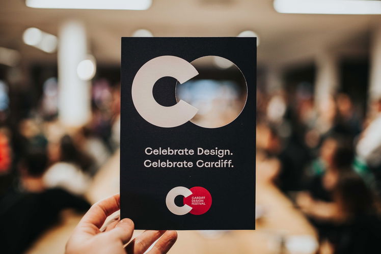A hand holds a dark card with bold white text reading “Celebrate Design. Celebrate Cardiff.” amid a blurred indoor crowd; the card features a circular logo and a red sticker for Cardiff Design Festival.