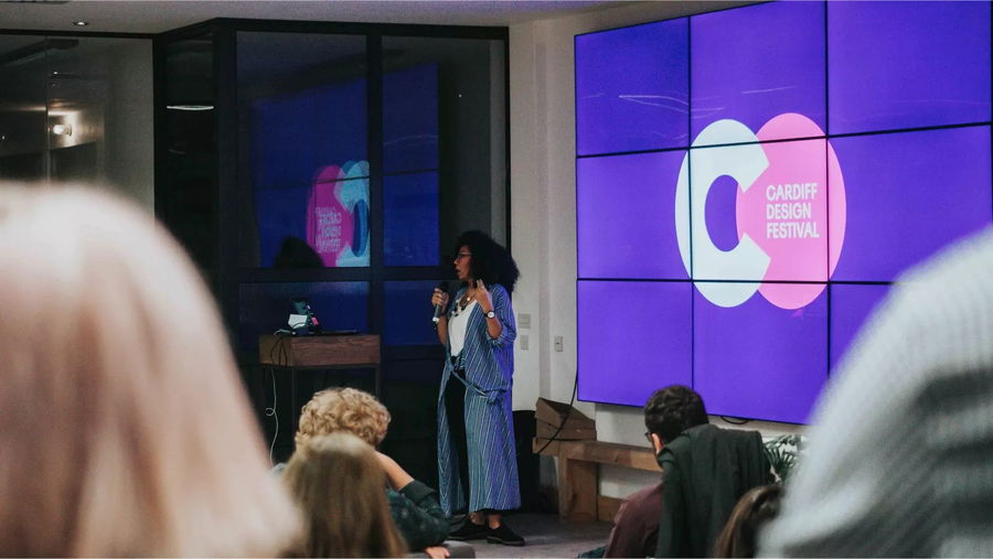 A woman speaks into a microphone in front of a large screen displaying the Cardiff Design Festival logo, as an audience listens in a modern indoor setting.