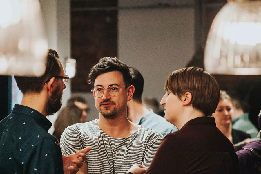 Three people are having a conversation in a busy indoor setting. The person in the centre is wearing glasses and a striped shirt, while the other two face him, with several blurred figures visible in the background.