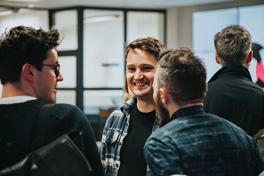 Three people stand talking indoors near an office window. The person in the middle is smiling, whilst the others face him, engaged in conversation. The background shows blurred office spaces and another person facing away.