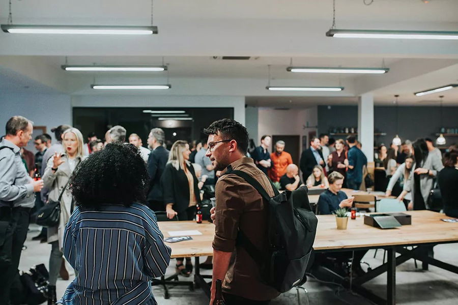 A large group of people socialise in a modern, well-lit office or event space. Some stand and chat, while others sit at tables with laptops and drinks. The atmosphere appears lively and professional.