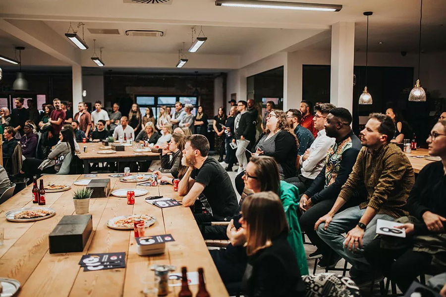 A large group of people sits and stands around long wooden tables, listening attentively to a speaker out of frame. Pizza, drinks, and papers are on the tables in a modern, well-lit room.