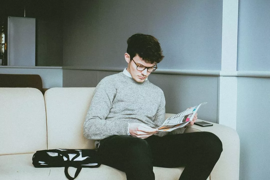 A young man wearing glasses and a grey jumper sits on a beige sofa reading a magazine, perhaps studying for University to Blue Stag exams. A black bag and mobile rest beside him. The background features pale walls and minimal decor.